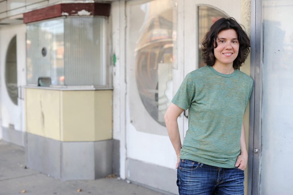 Woman with shoulder length brown hair and green t-shirt leaning against a white door.