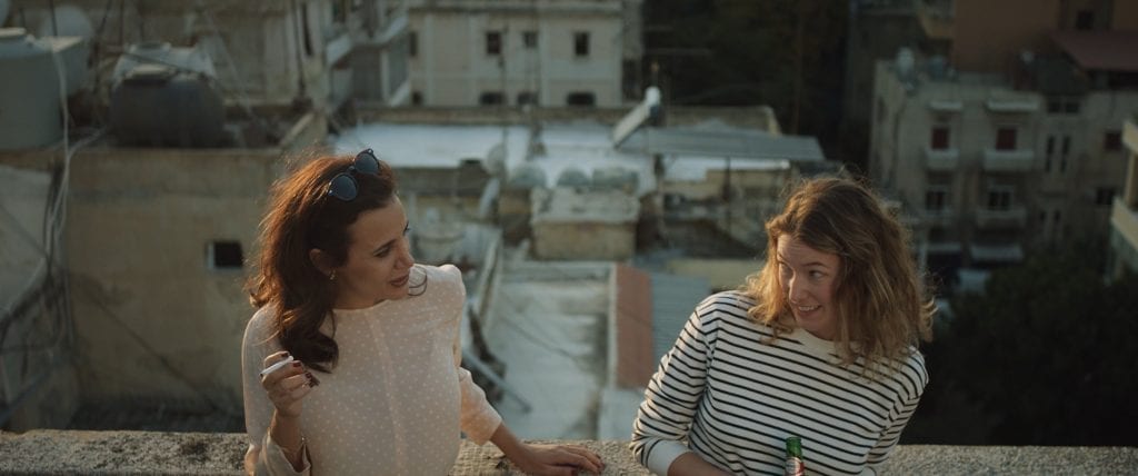 Two women looking playfully at each other with short urban buildings off the balcony behind them