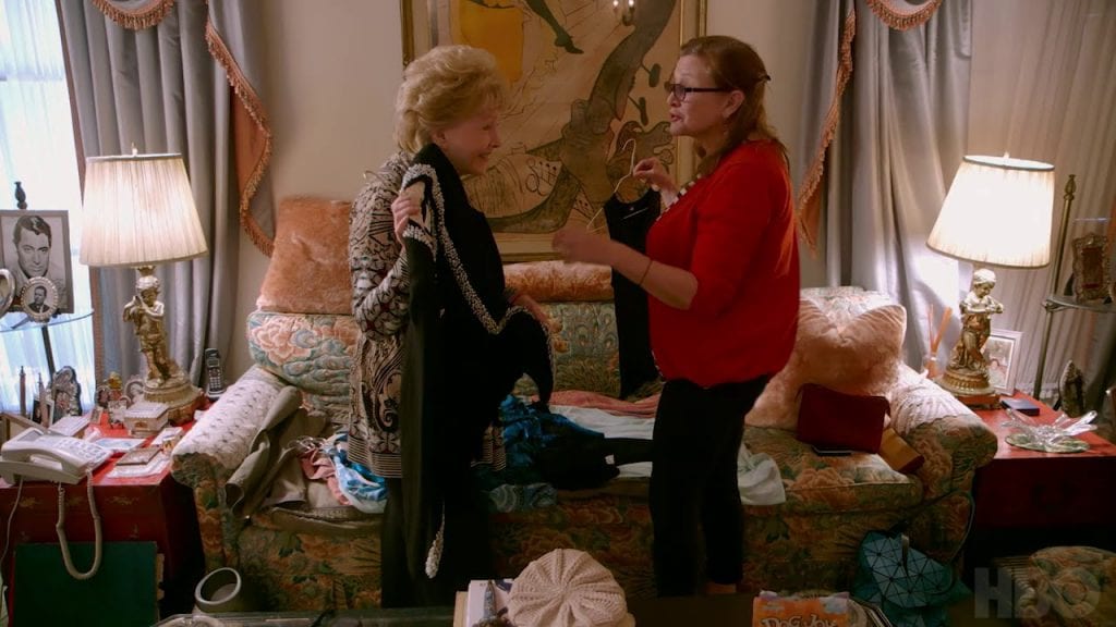 An elderly mother and middle aged daughter standing over a couch, holding clothes up to each other