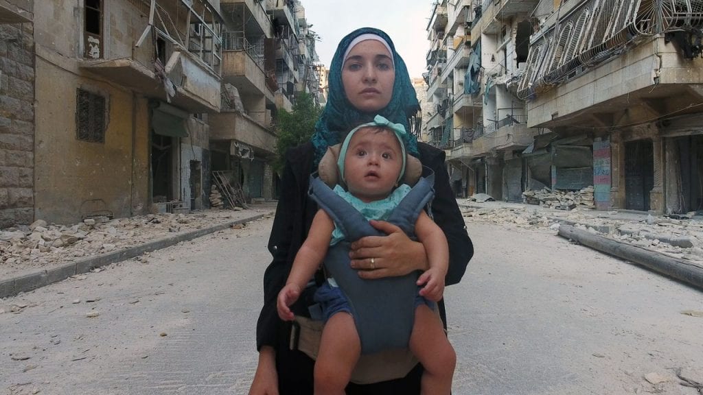 A woman standing in an empty street, carrying a baby in a front carrier.