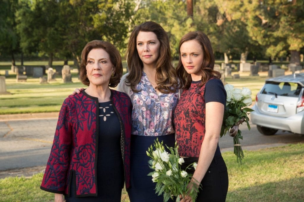 three women, a grandmother, daughter, and granddaughter, standing next to each other in a cemetery looking off screen.