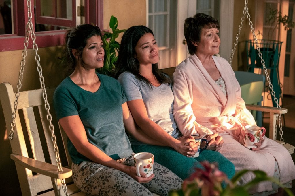 Three women, a grandmother, daughter, and granddaughter, sit on a swinging front porch bench in pajamas, holding hands and looking offscreen.