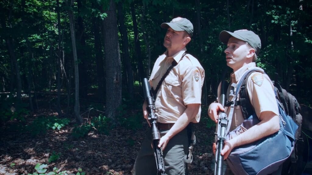 Lyle (Ben Guenther) and Victor (Christ Young) stand next to each other in the woods wearing khaki park ranger uniforms and holding shotguns. 