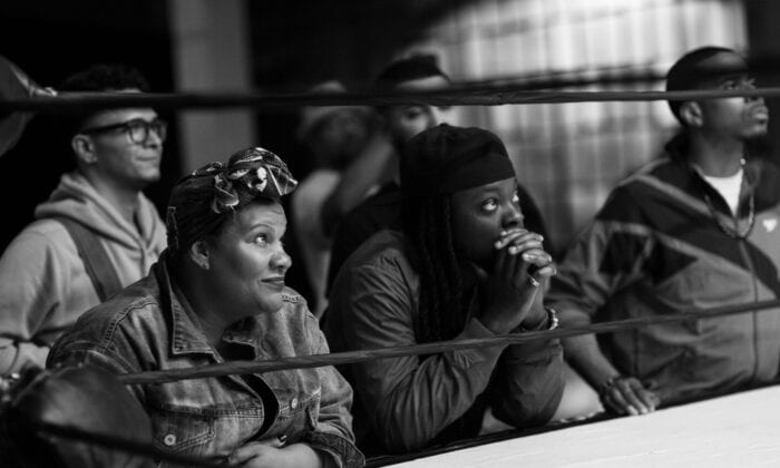 A balck-and-white still from The Forty-Year-Old Version where some of the characters are sitting near the edge of a boxing ring, watching something. 