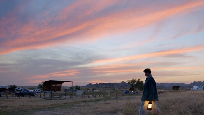 A still from Nomadland where a character walks through a field during a sunset, alone, carrying a lantern. 