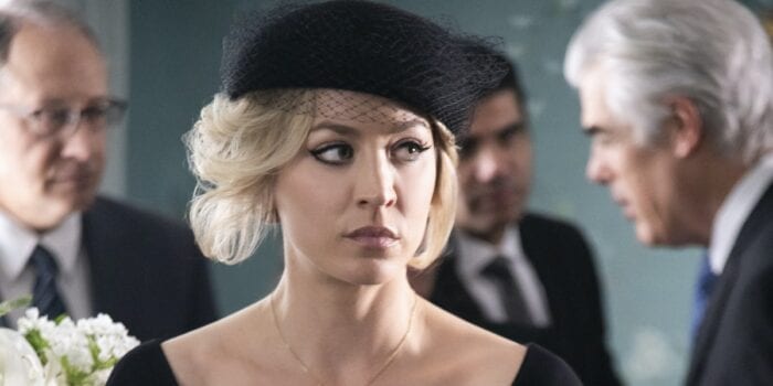 A close up of Cassie dressed in funeral garb (including a vintage-style hat with netting on it) looking off to the side while other funeral attendees stalk behind her.