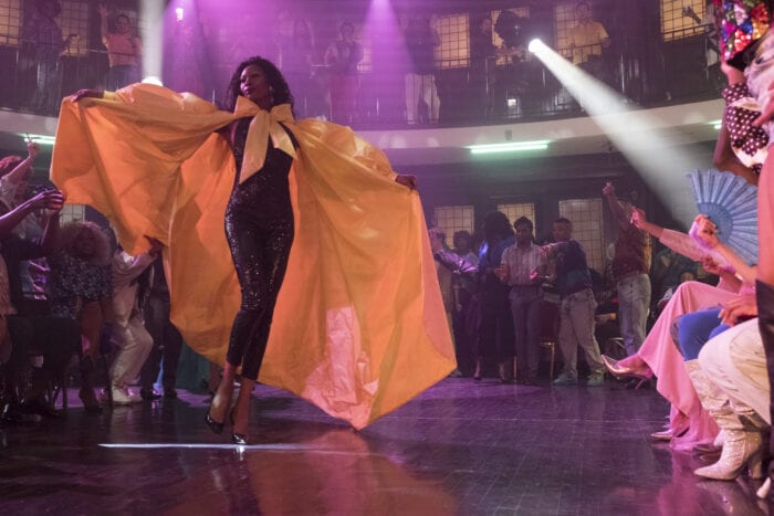 A still from Pose, featuring a character dressed in a shiny black jumpsuit and orange cape in a room of happy, partying people. 