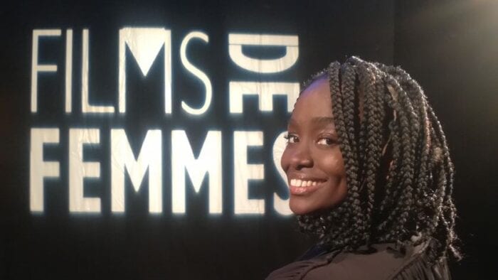 An image of Aïssa Maïga sitting in a black, folding director's chair with "films de femmes" written on the back. She is a black room with a light up sign of "films de femmes."