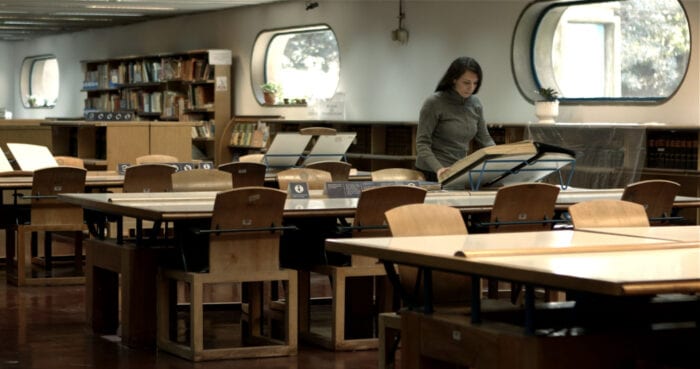 Mara stands at a table over a large book on a stand in a library, doing research for her film. 
