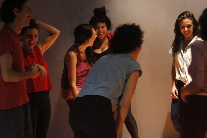 Avila stands with a group of dancers in a dimly lit set, smiling and laughing between takes. 