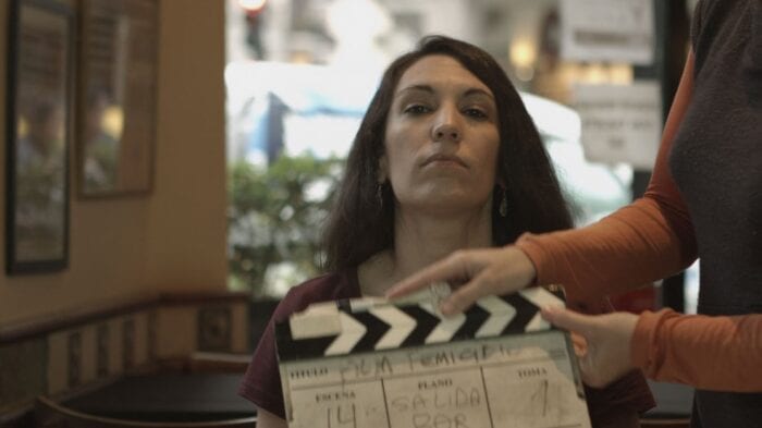 Mara stared down the camera while sitting at a table, a man holding a clapboard in front of her. 
