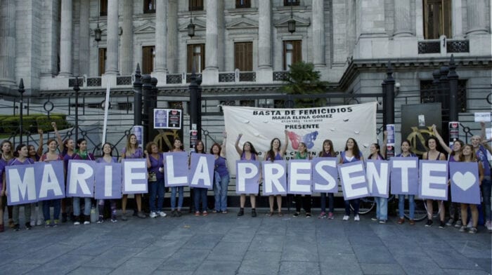 Women stand in a line outside of what appears to be a government building, holding up large lettered signs that spell "Mariela Presente."