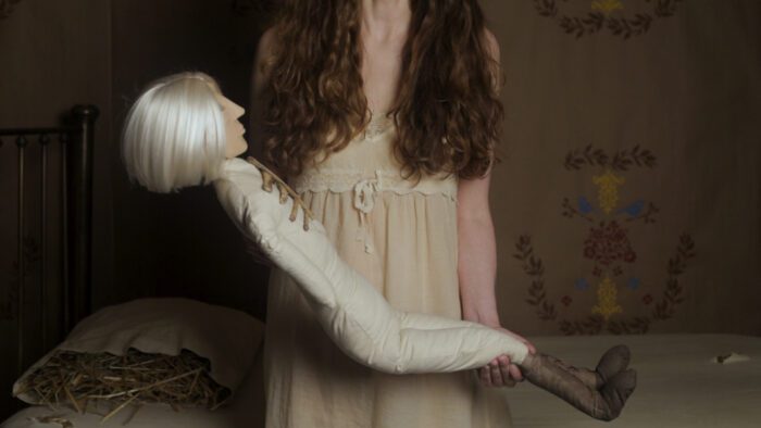 Still of a woman viewed from the neck down, holding a medium-large stuffed doll with white hair and a visible rib cage. They are in an old-timey yet simple room with a bed and pillow full of straw.