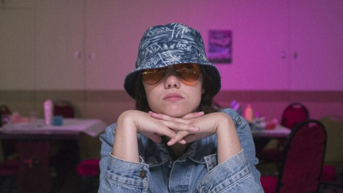 An actor sits with their head resting on their laced fingers in a very pink-toned room. They are wearing yellow-tinted aviator glasses, a blue, beachy bucket hat, and a denim shirt.
