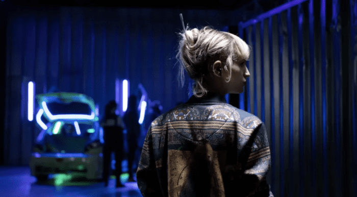 Alexia is standing in a blue neon-lit warehouse, wearing an ornate bomber jacket and her blond hair up in a twist.
