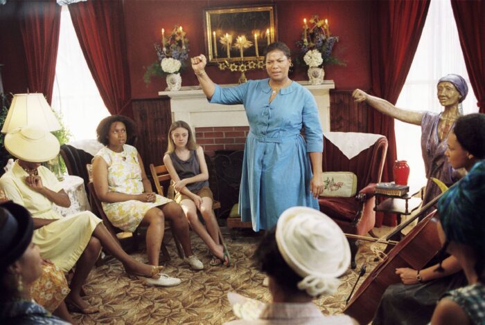 August stands in a bright blue dress with her fist raised in the center of a room of seated black women in church clothes. Lily is also there, wearing much less formal clothes.