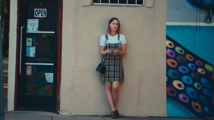 Lady Bird stands against a nondescript street corner, wearing a plaid pinafore and white t-shirt.