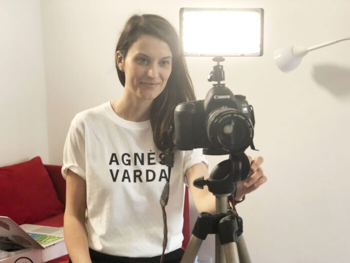 Elena sits behind a camera, with a small smile and an agnes varda shirt