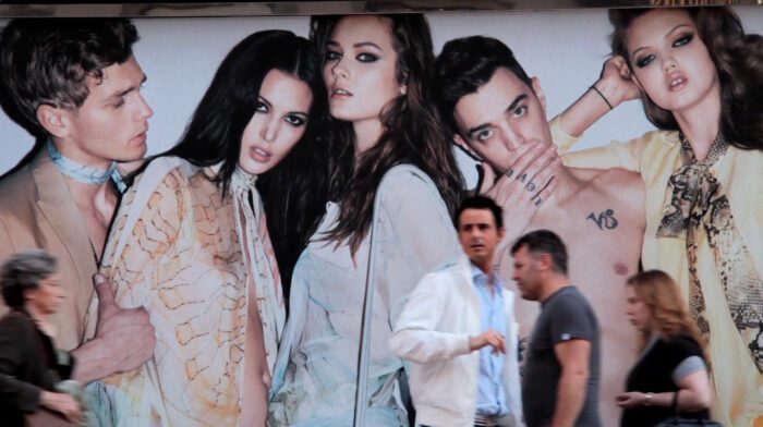 A still of blurred people walking in front of a large advertisement featuring thin, white models