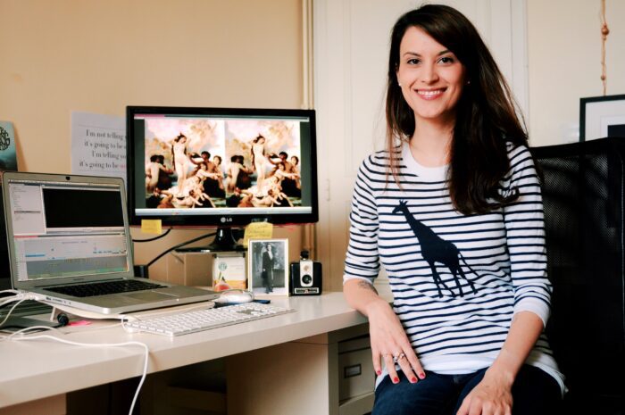 Elena sits, smiling, next to a computer with video editing software open on it.
