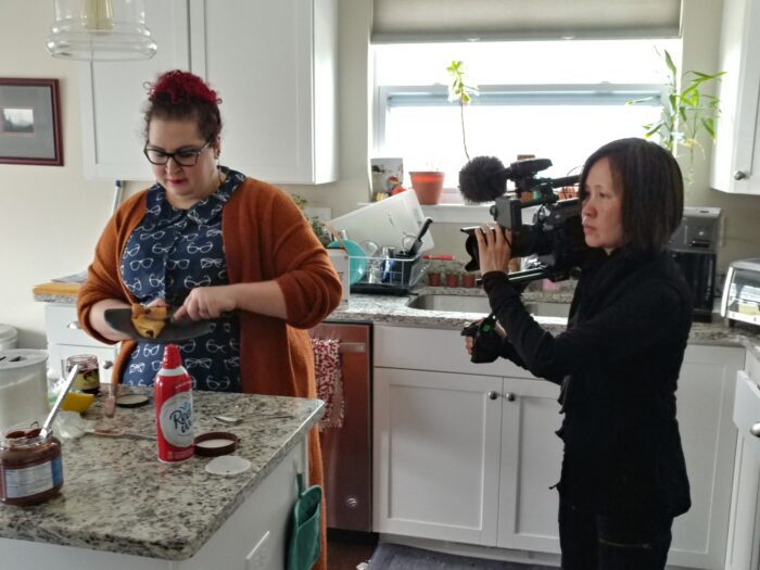 A woman in all black points a camera at another woman in a printed shirt who's making lunch in her kitchen. 