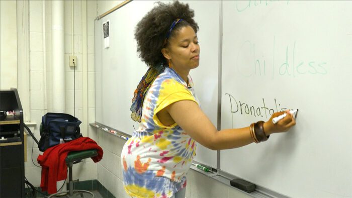 Kimya, in a tie-dye shirt, writes the word "pronatalism" on a white board. 