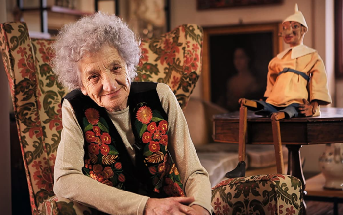 Cecilia Mangini, and old woman, leans into the camera and smiles with a puppet on a table behind her. 