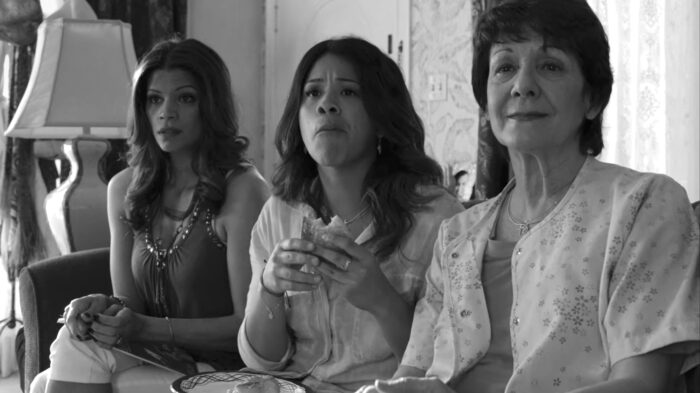 A black-and-white still from Jane the Virgin where three women are sat together on a couch, the middle one in the process of eating a sandwich.
